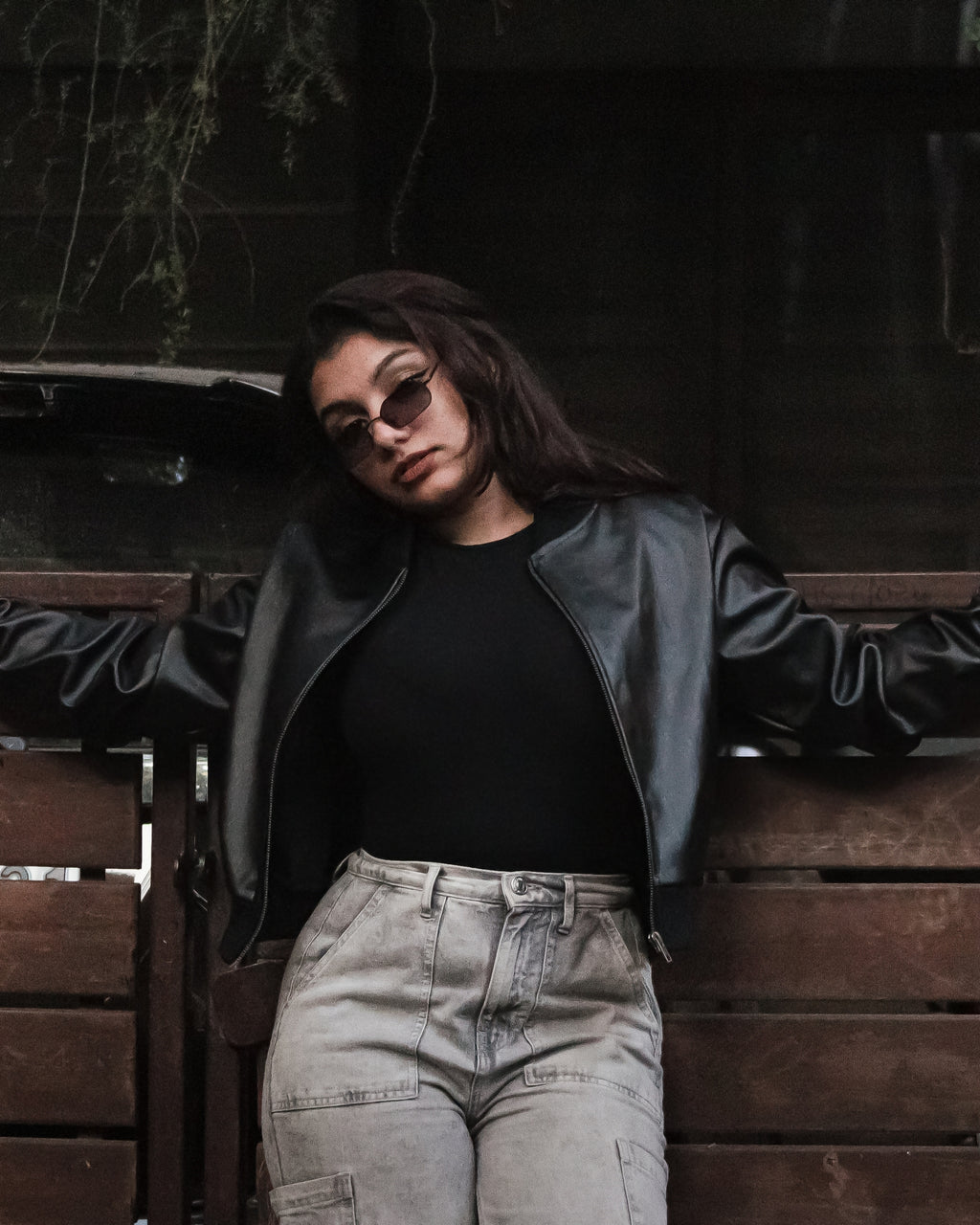 Person wearing a black leather jacket and sunglasses standing against a wooden wall.