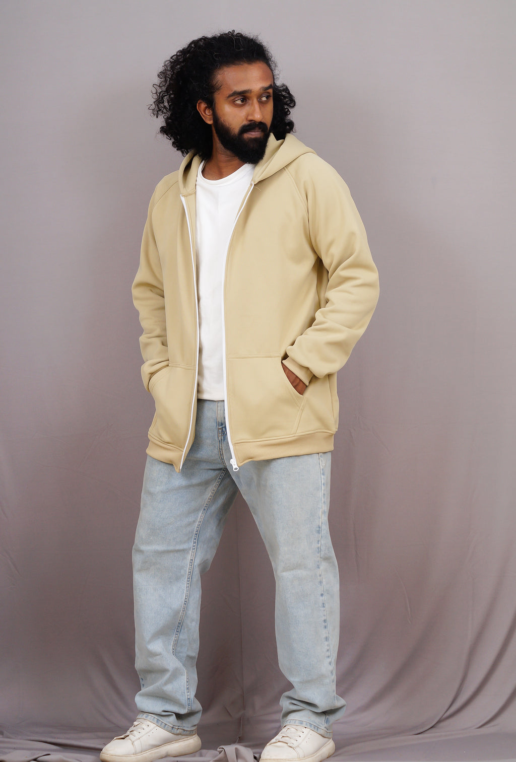 Man wearing a beige hoodie and light blue jeans against a gray background