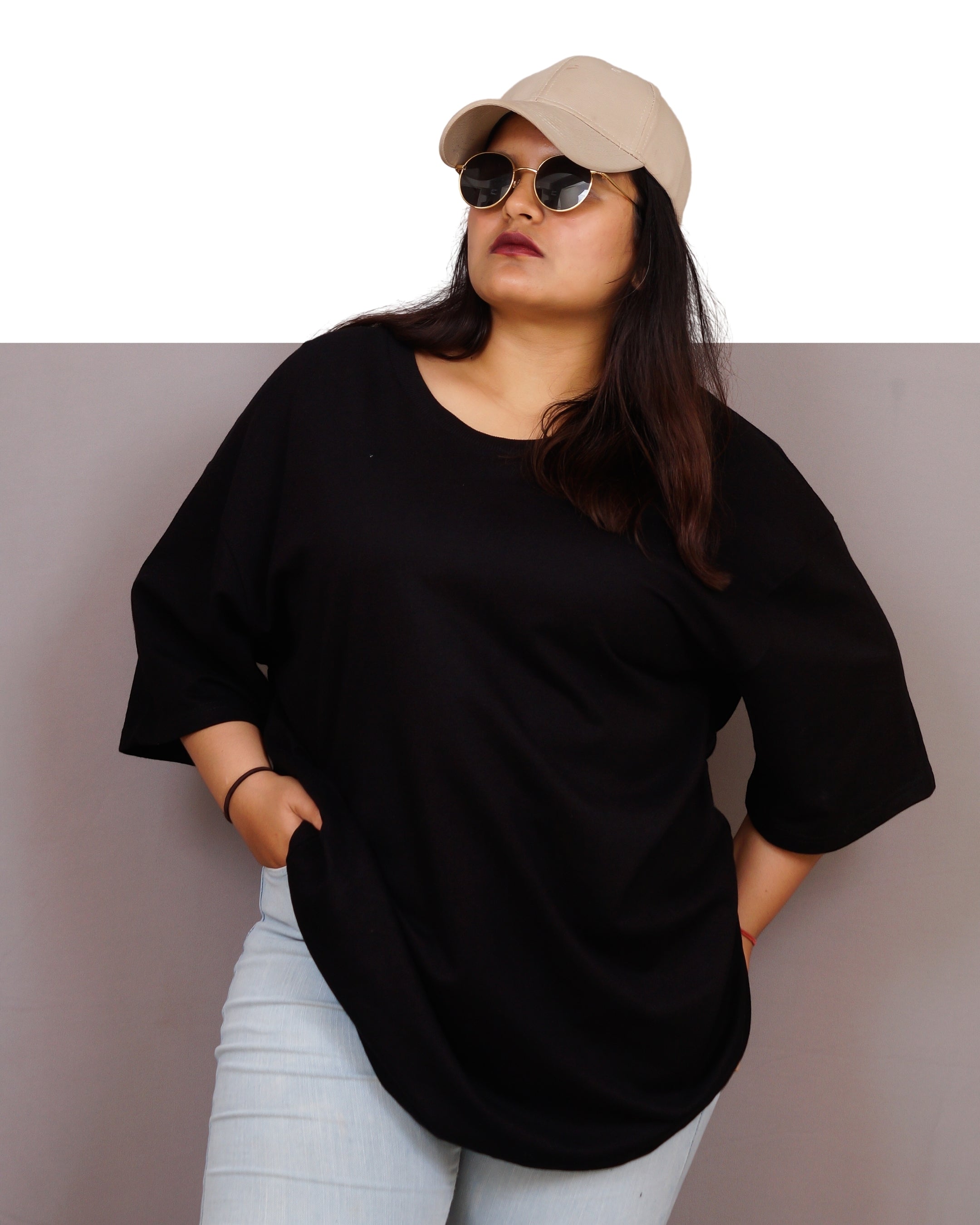 Person wearing a black oversized shirt, light blue jeans, and a beige cap with sunglasses on a plain background