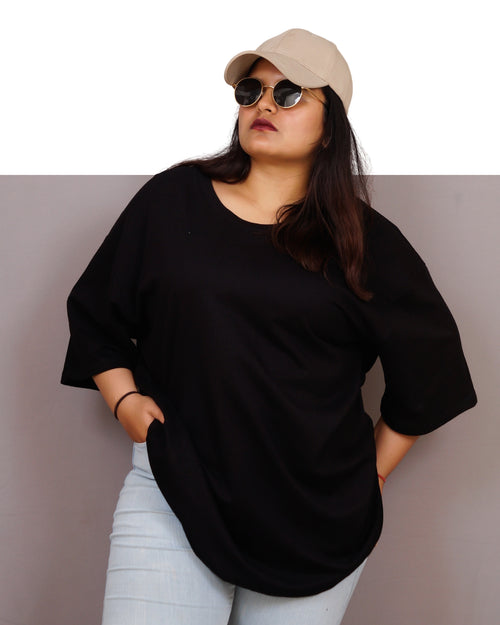 Person wearing a black oversized shirt, light blue jeans, and a beige cap with sunglasses on a plain background