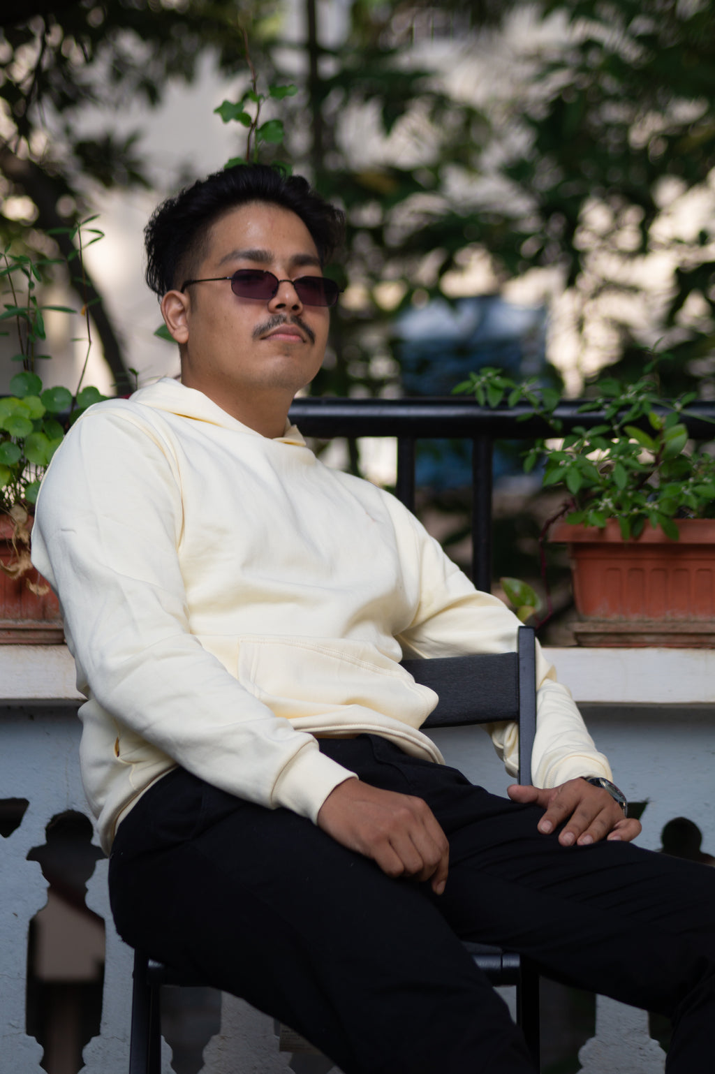 Person wearing sunglasses and a white sweater sitting outdoors with plants in the background