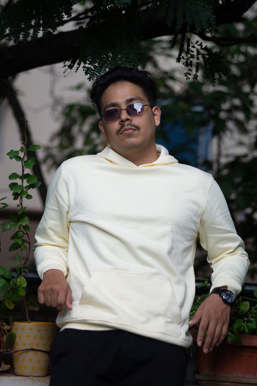 Person wearing a white hoodie and sunglasses sitting outdoors with plants in the background