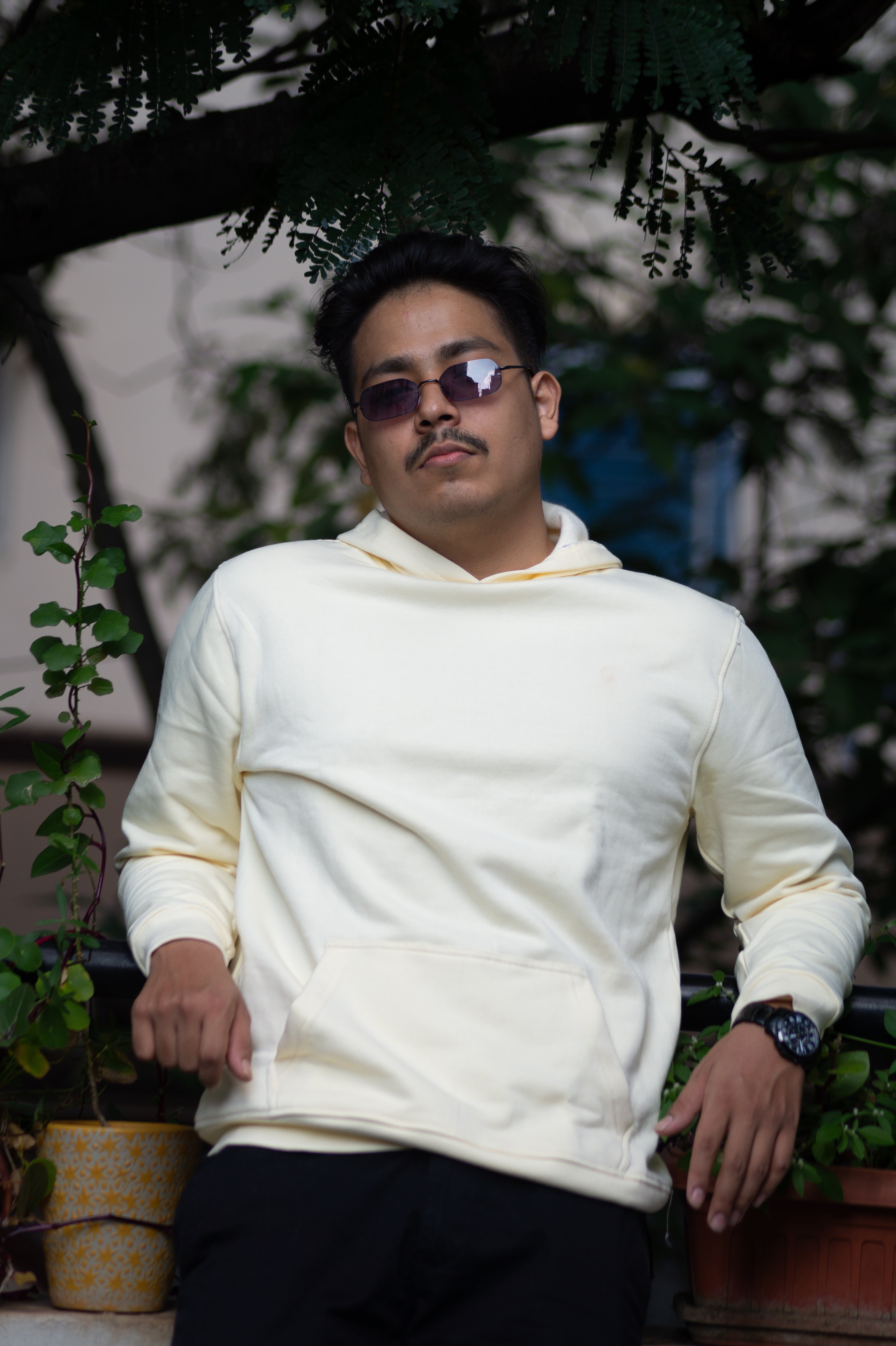 Person wearing a white hoodie and sunglasses sitting outdoors with plants in the background