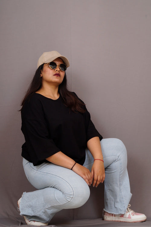 Woman wearing a black top, light blue jeans, beige cap, and sunglasses against a plain background