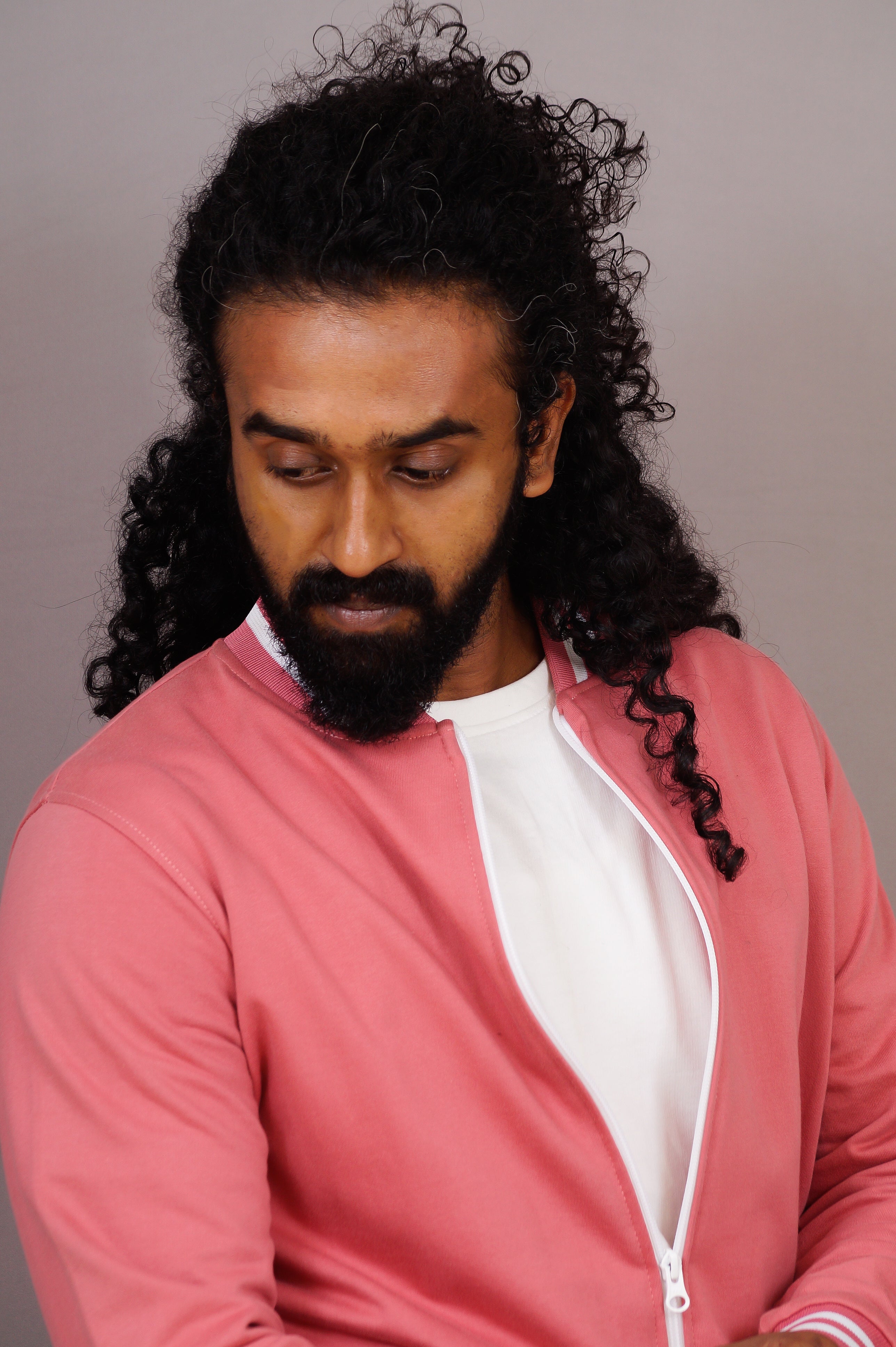 Man with long hair and beard wearing a pink jacket over a white shirt against a gray background