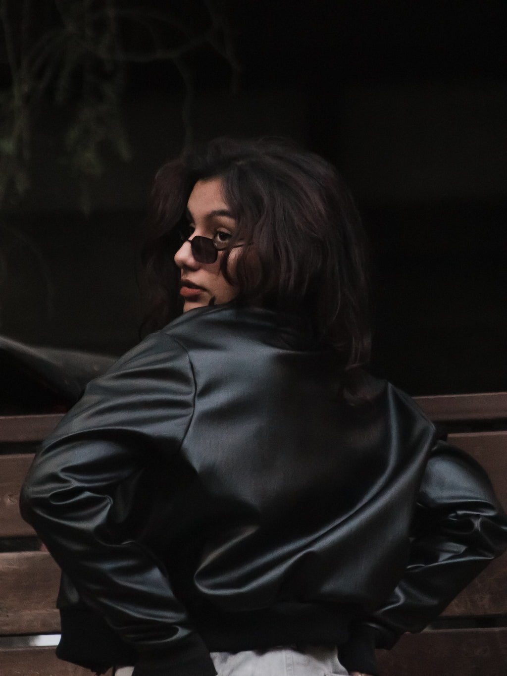 Person wearing a black leather jacket sitting on a bench with a dark background
