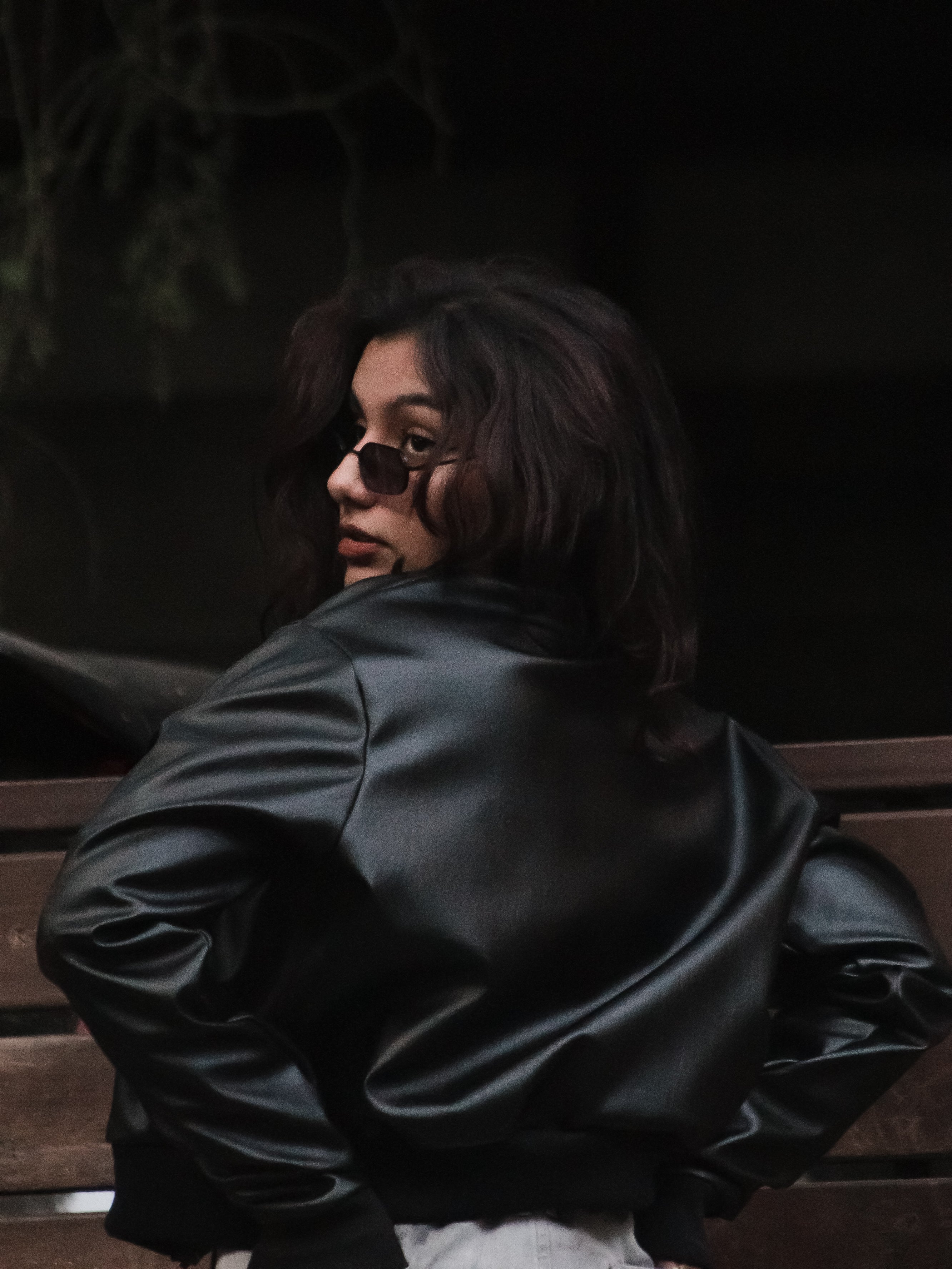 Person wearing a black leather jacket sitting on a bench with a dark background