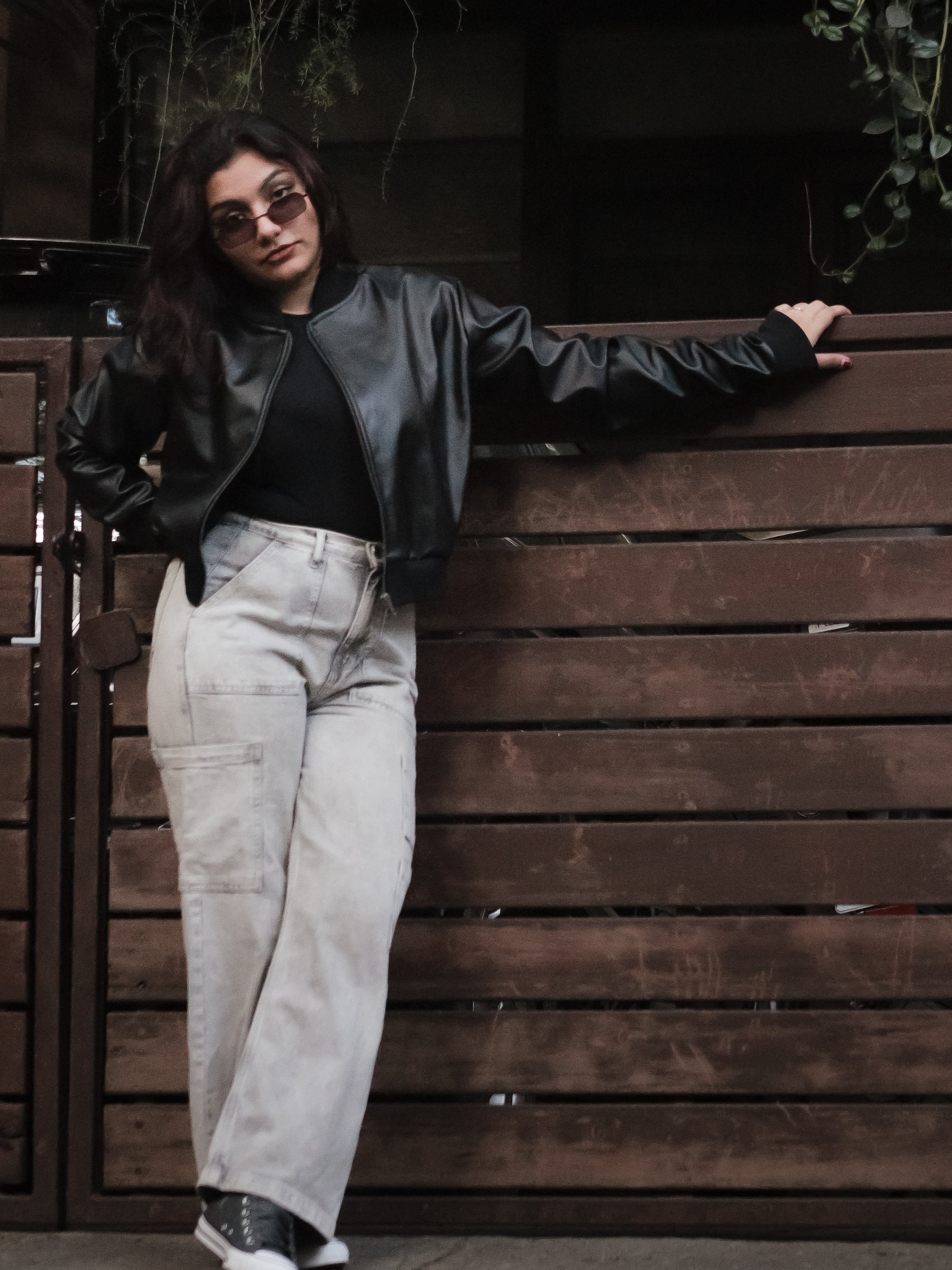 Person wearing a black leather jacket and light-colored pants leaning against a wooden wall.