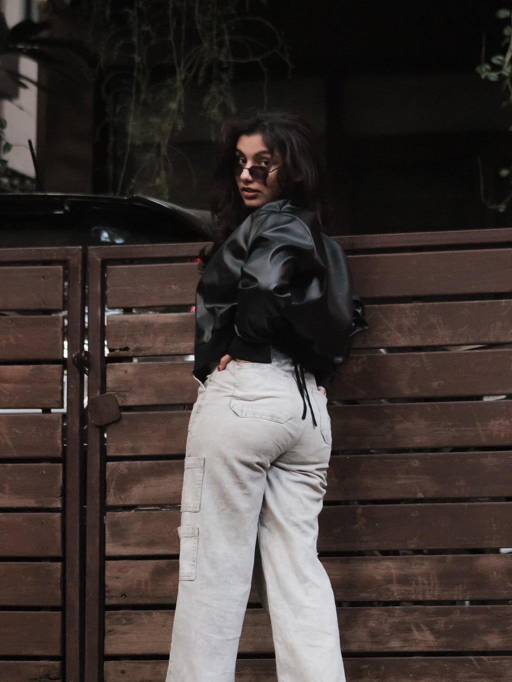 Person wearing a black jacket and light-colored pants leaning against a wooden wall.