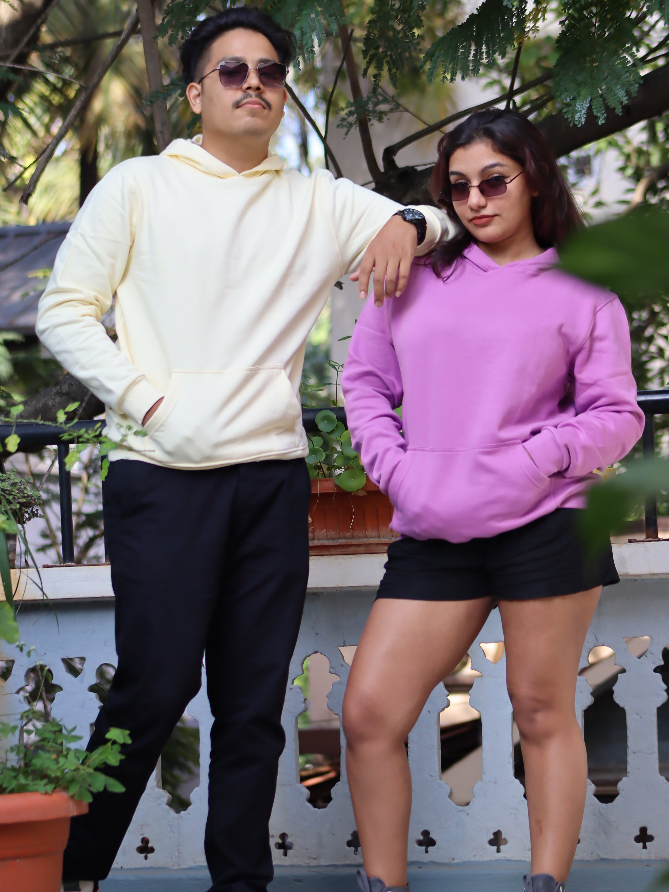 Two people wearing hoodies standing outdoors with plants in the background