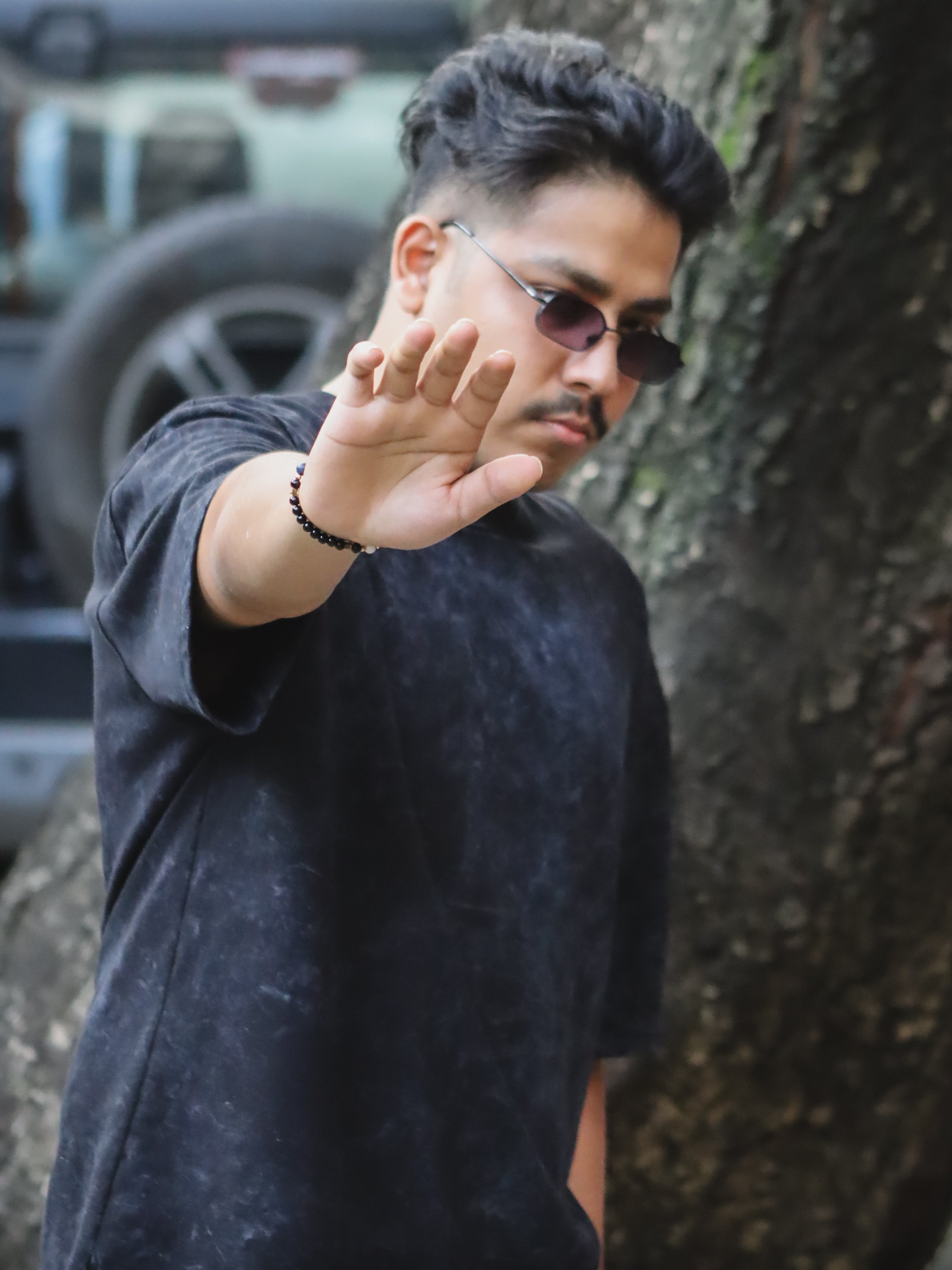 Man wearing sunglasses and a dark shirt, standing outdoors with a tree and vehicle in the background