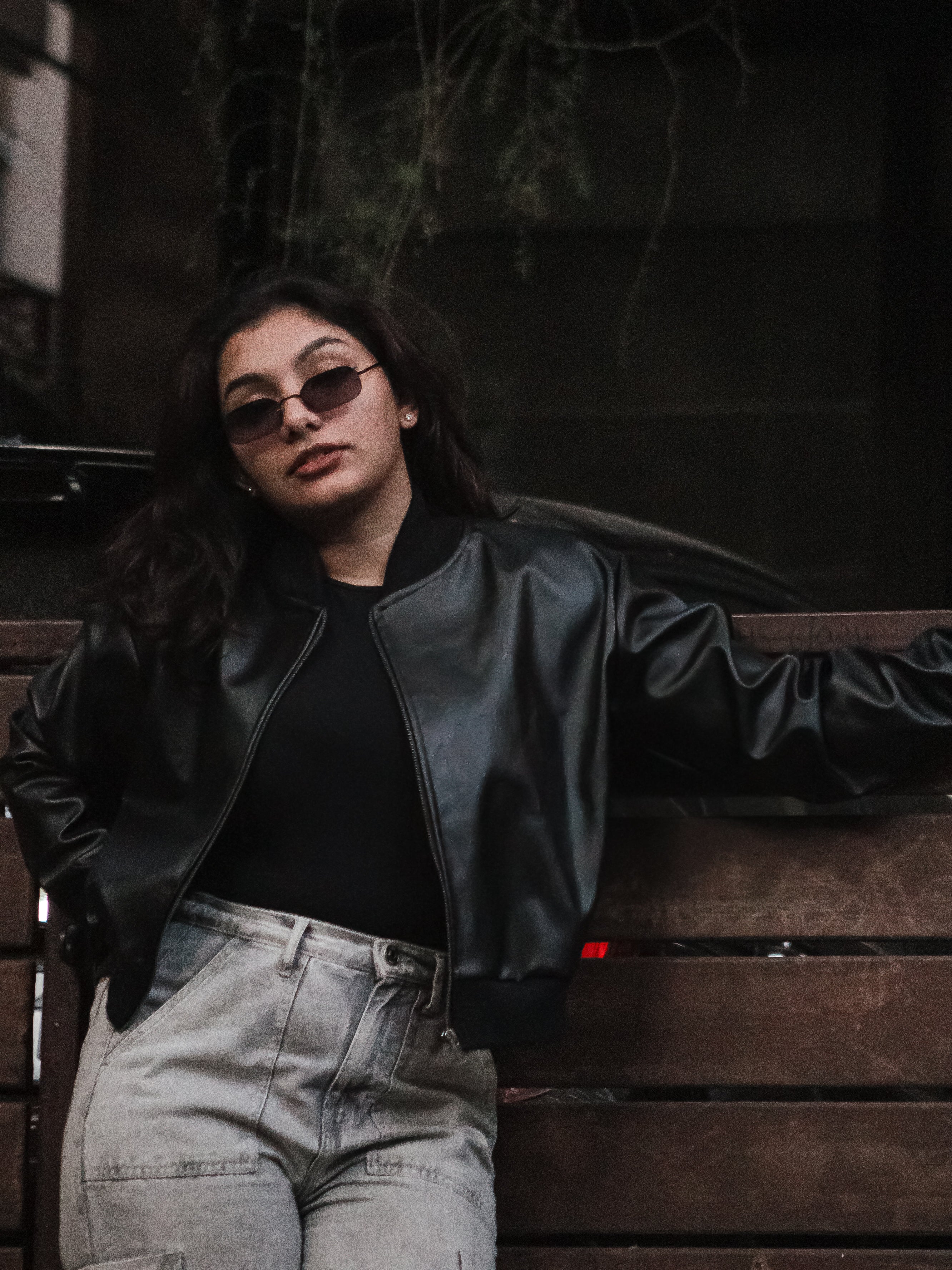 Person wearing a black leather jacket and sunglasses leaning against a dark wall.