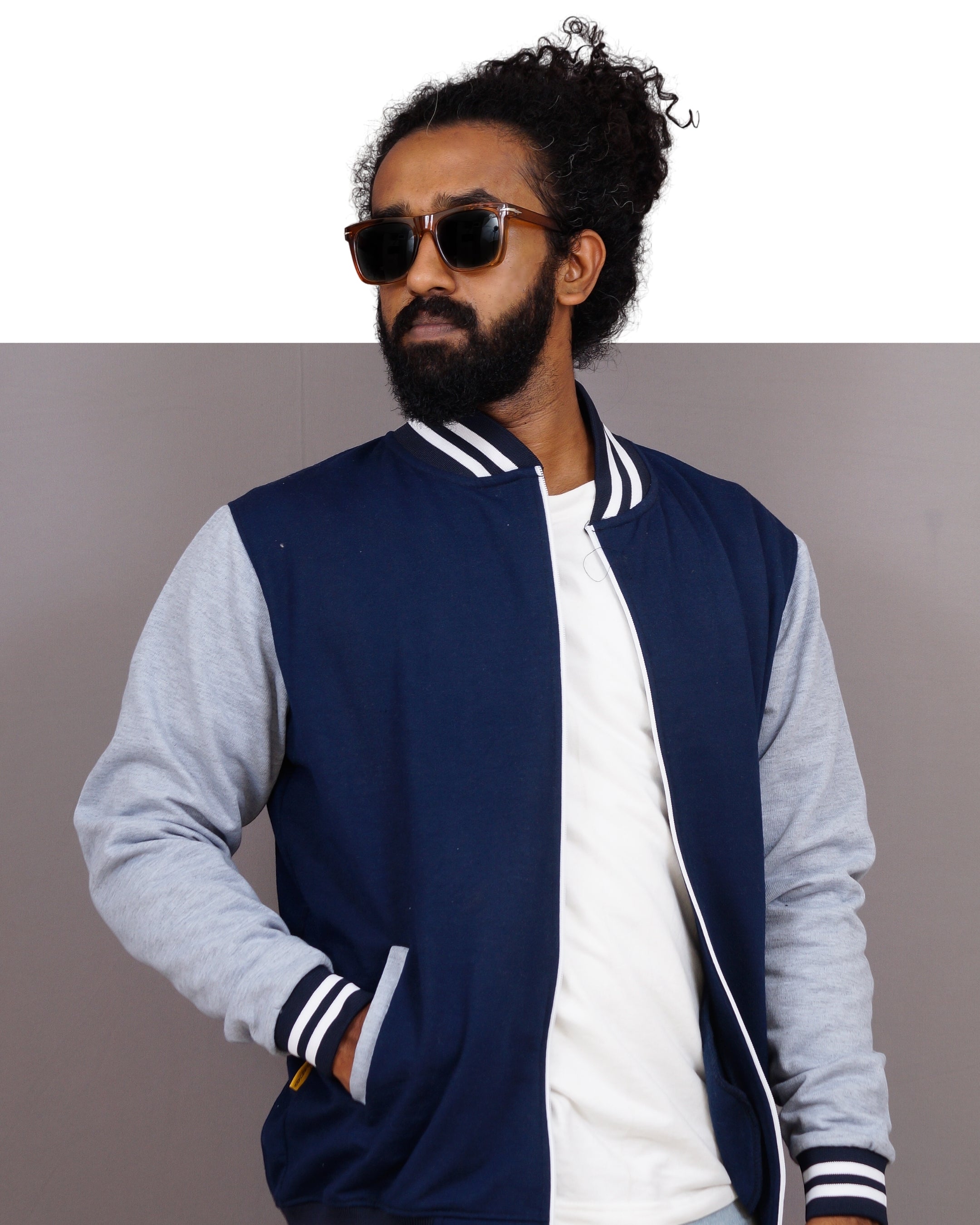 Man wearing a navy and gray varsity jacket with sunglasses on a plain background