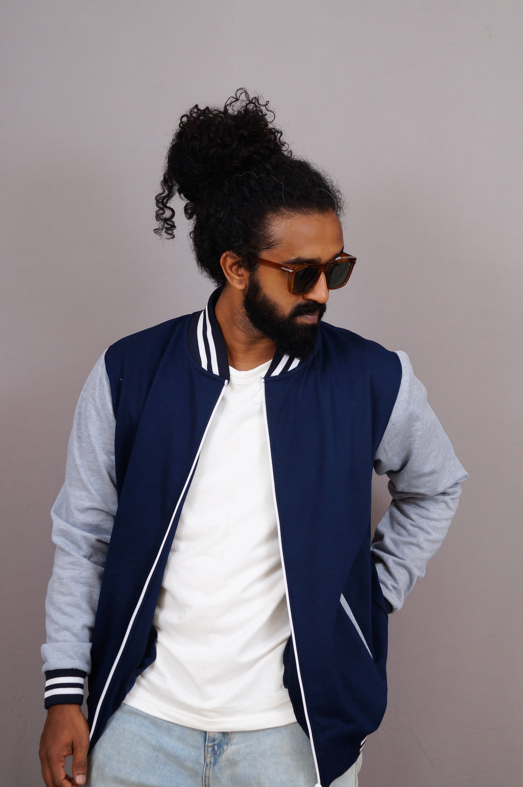 Man wearing a navy and gray varsity jacket with sunglasses and a white shirt against a plain background
