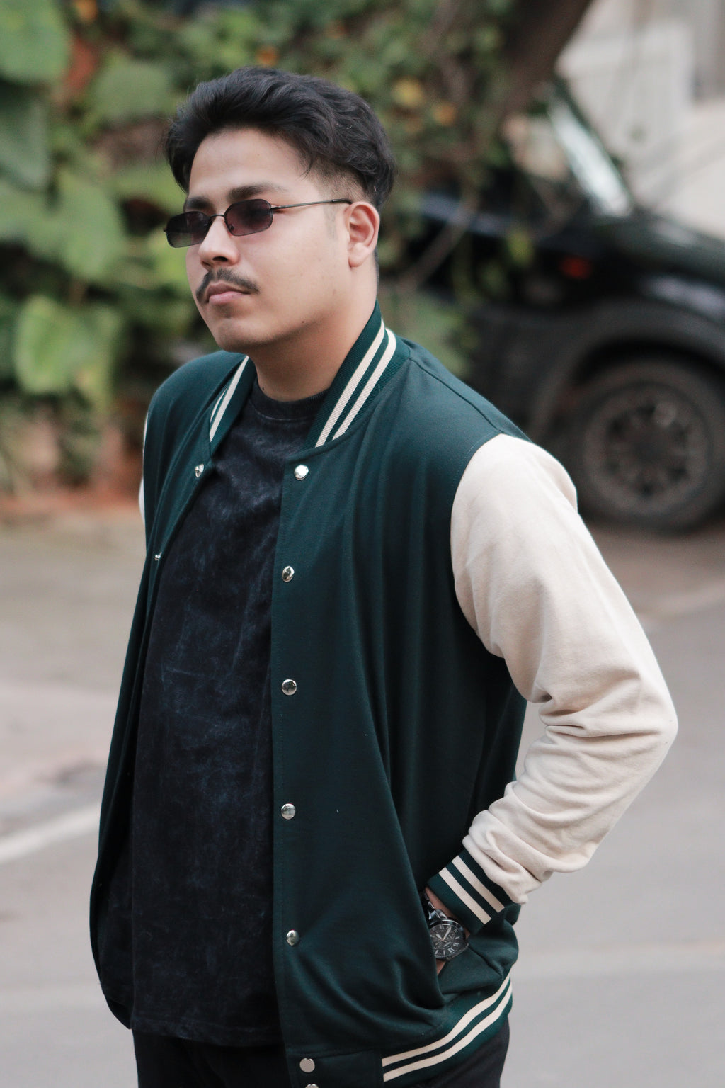 Man wearing a green varsity jacket with white sleeves and black sunglasses outdoors.
