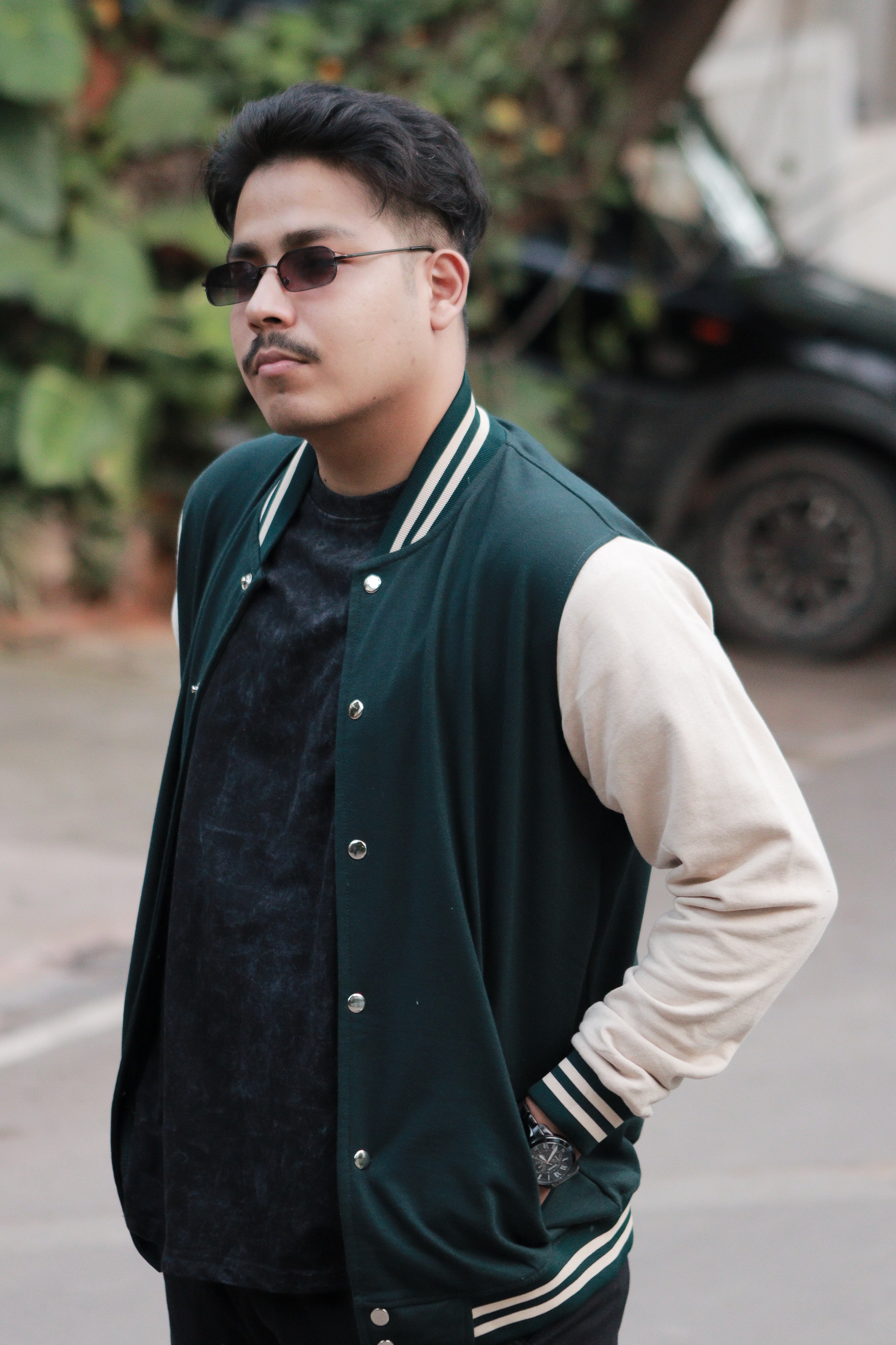 Man wearing a green varsity jacket with white sleeves and black sunglasses outdoors.