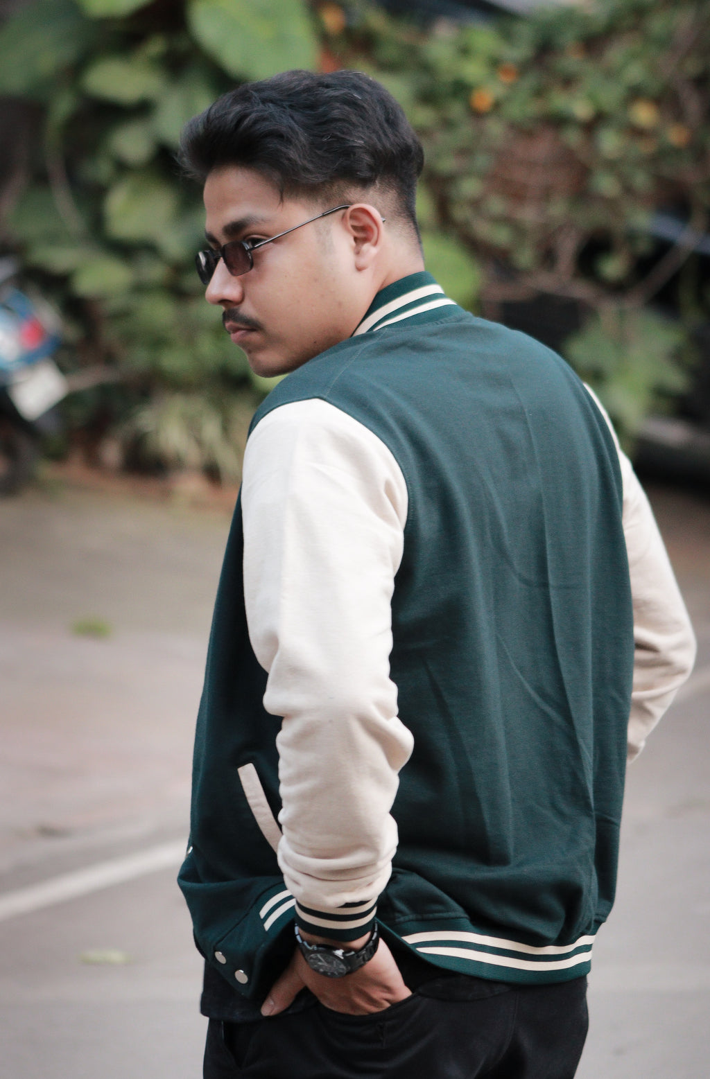 Man wearing a green and white varsity jacket with sunglasses and a beard, standing outdoors.