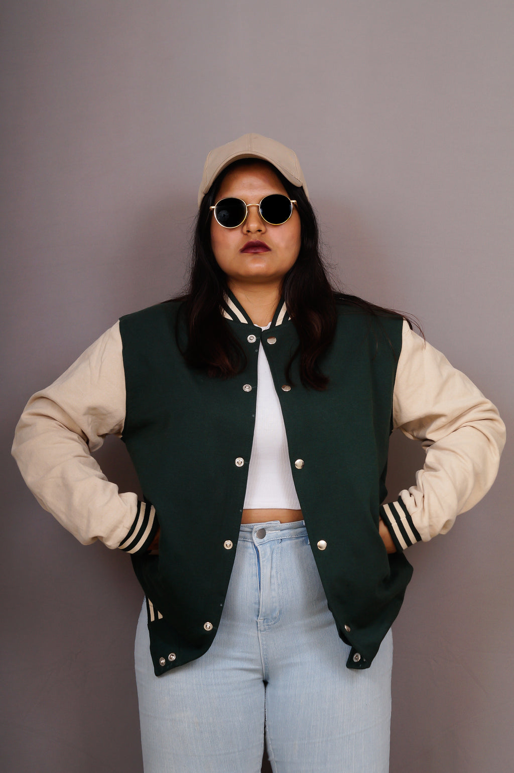 Person wearing a green varsity jacket over a beige sweater, light blue jeans, and sunglasses against a plain background.