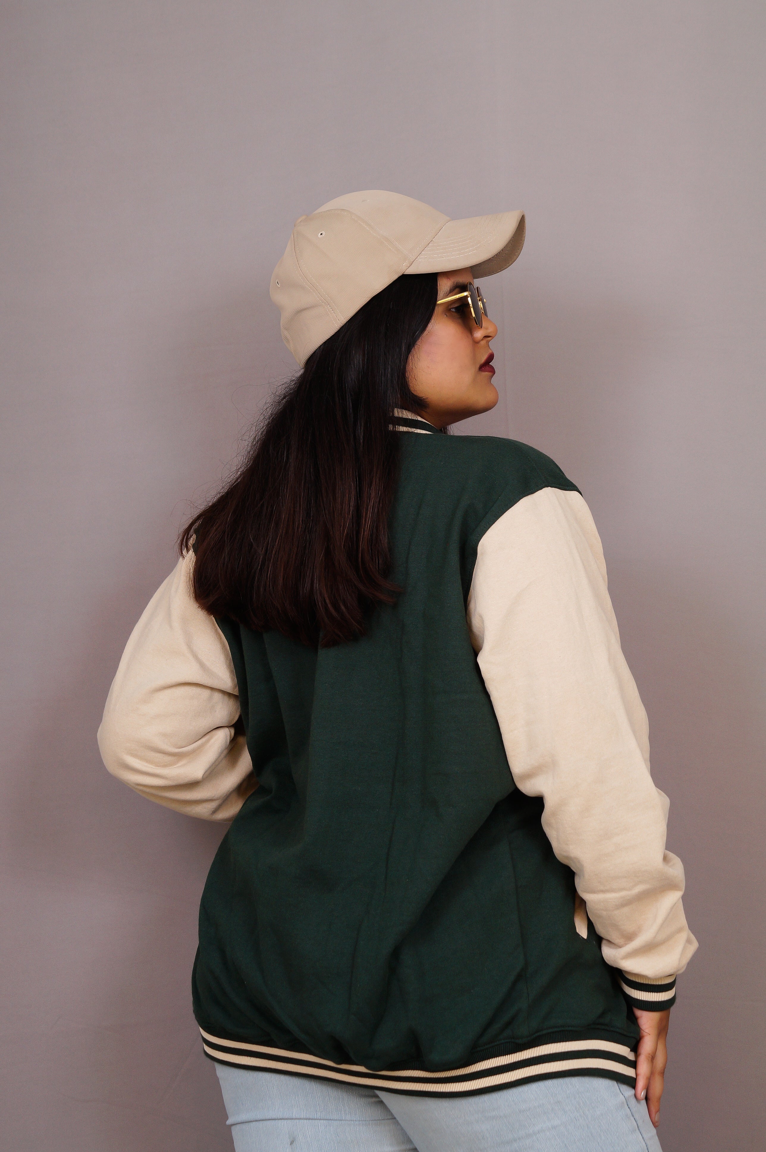 Person wearing a green and beige varsity jacket with a beige cap and sunglasses on a plain background