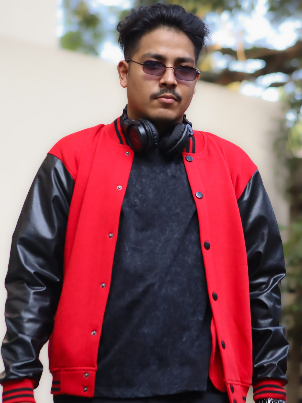 Man wearing a red and black varsity jacket with headphones around his neck, standing outdoors.