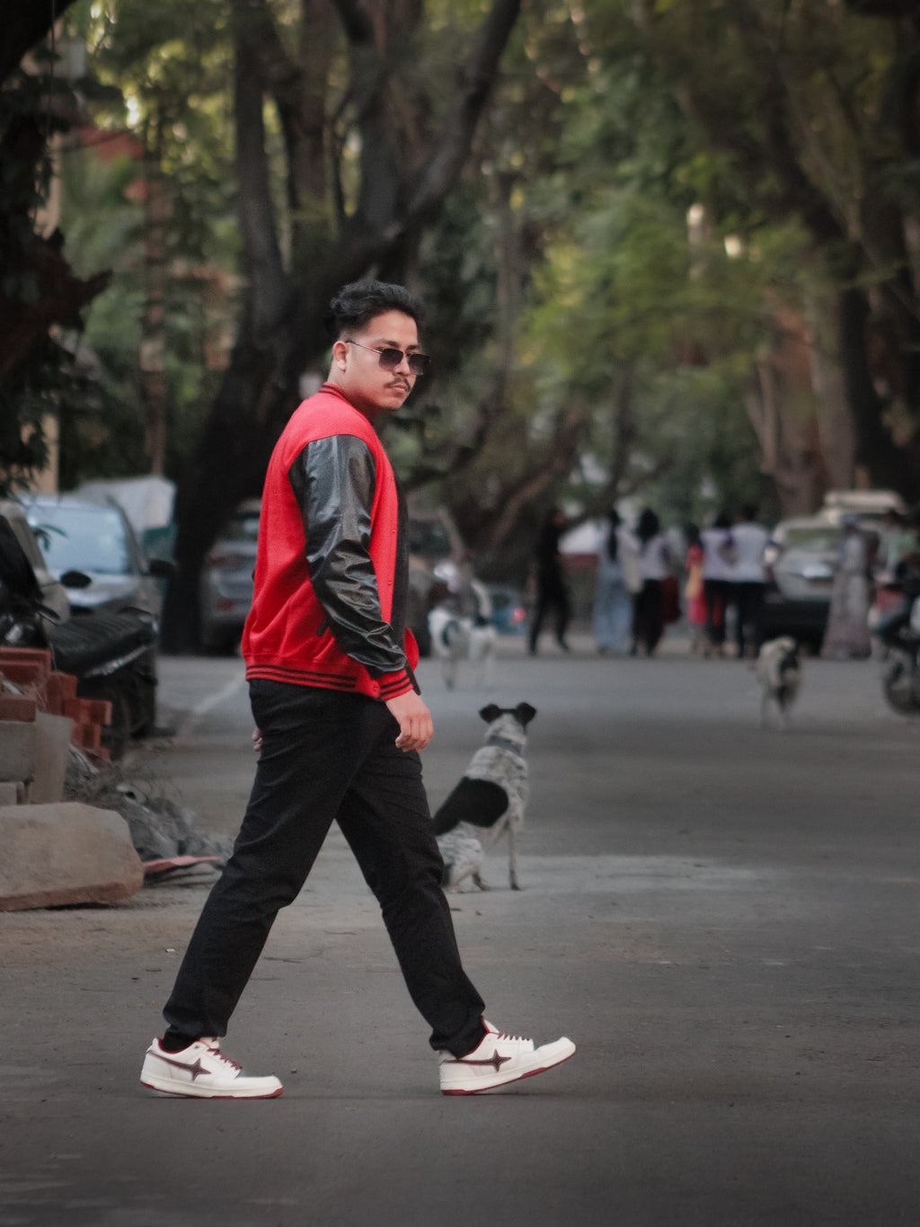Man walking on a street wearing a red and black jacket, sunglasses, and black pants.