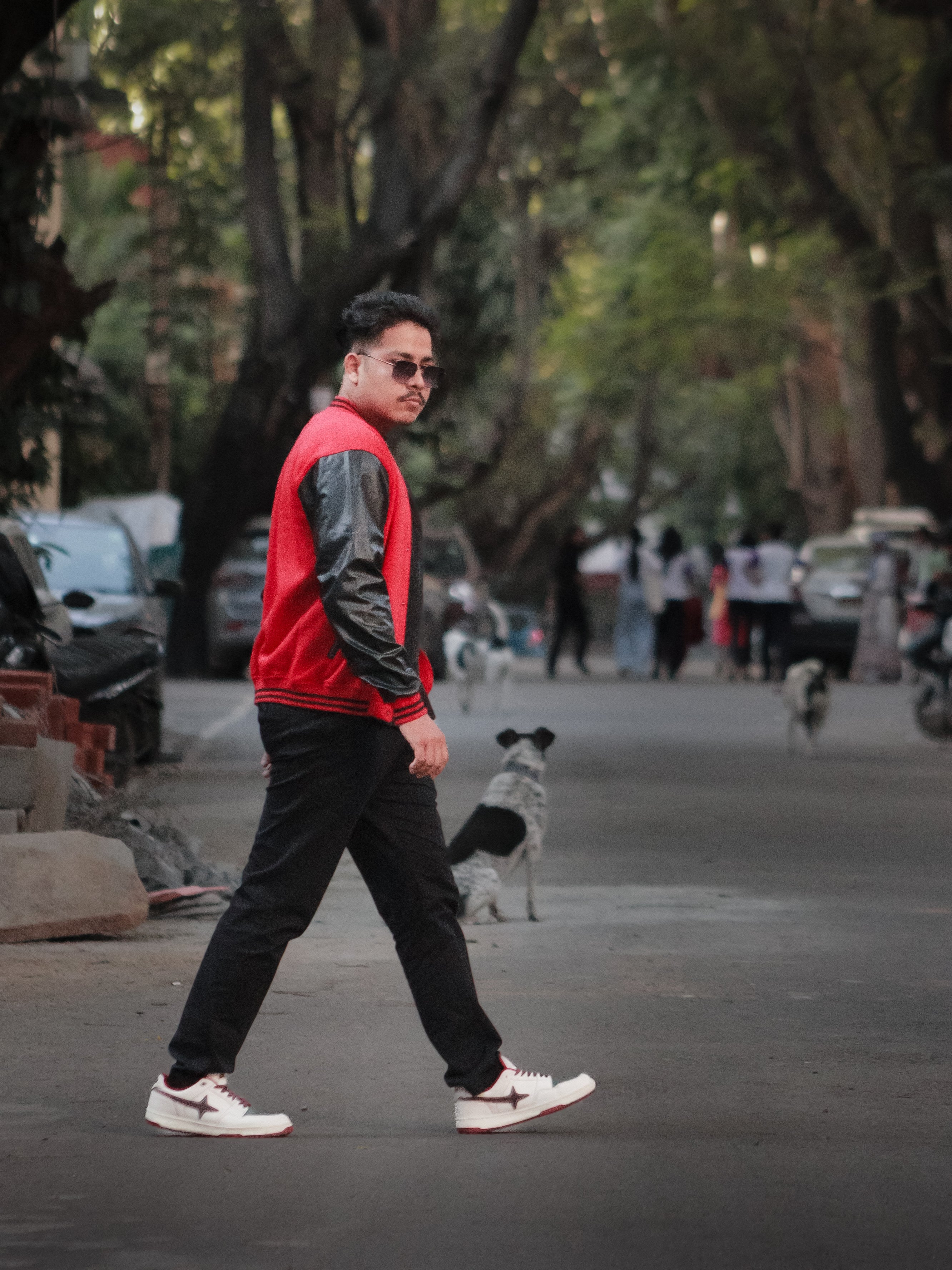 Man walking on a street wearing a red and black jacket, sunglasses, and black pants.