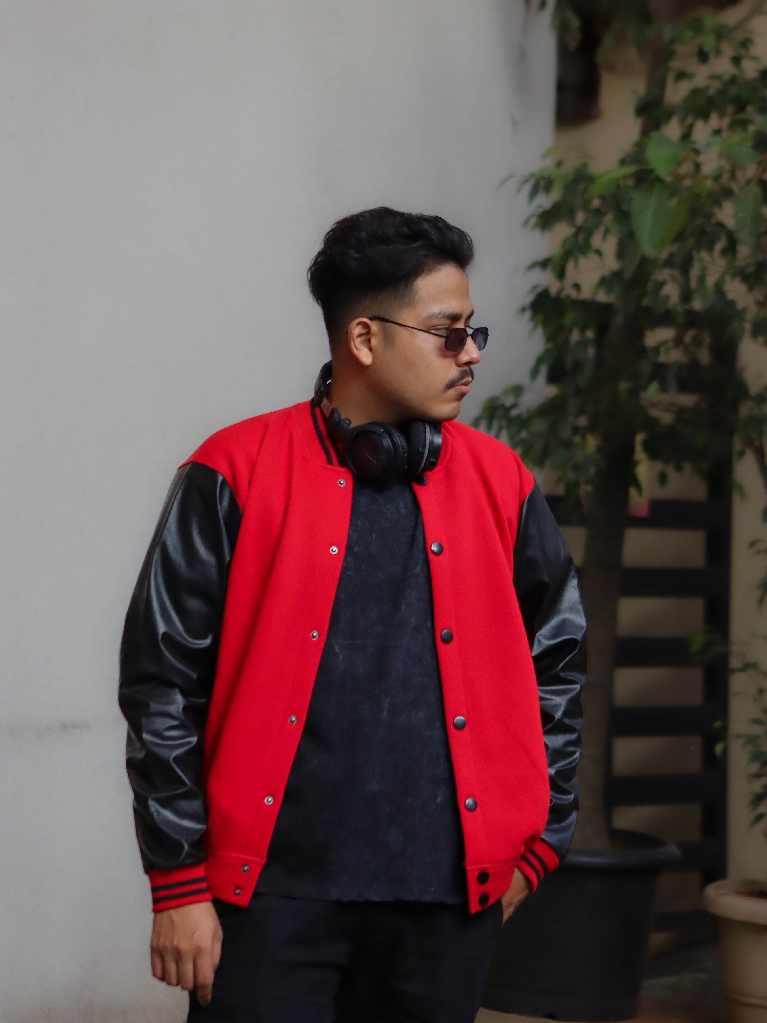 Man wearing a red and black varsity jacket with headphones around his neck, standing against a neutral wall.