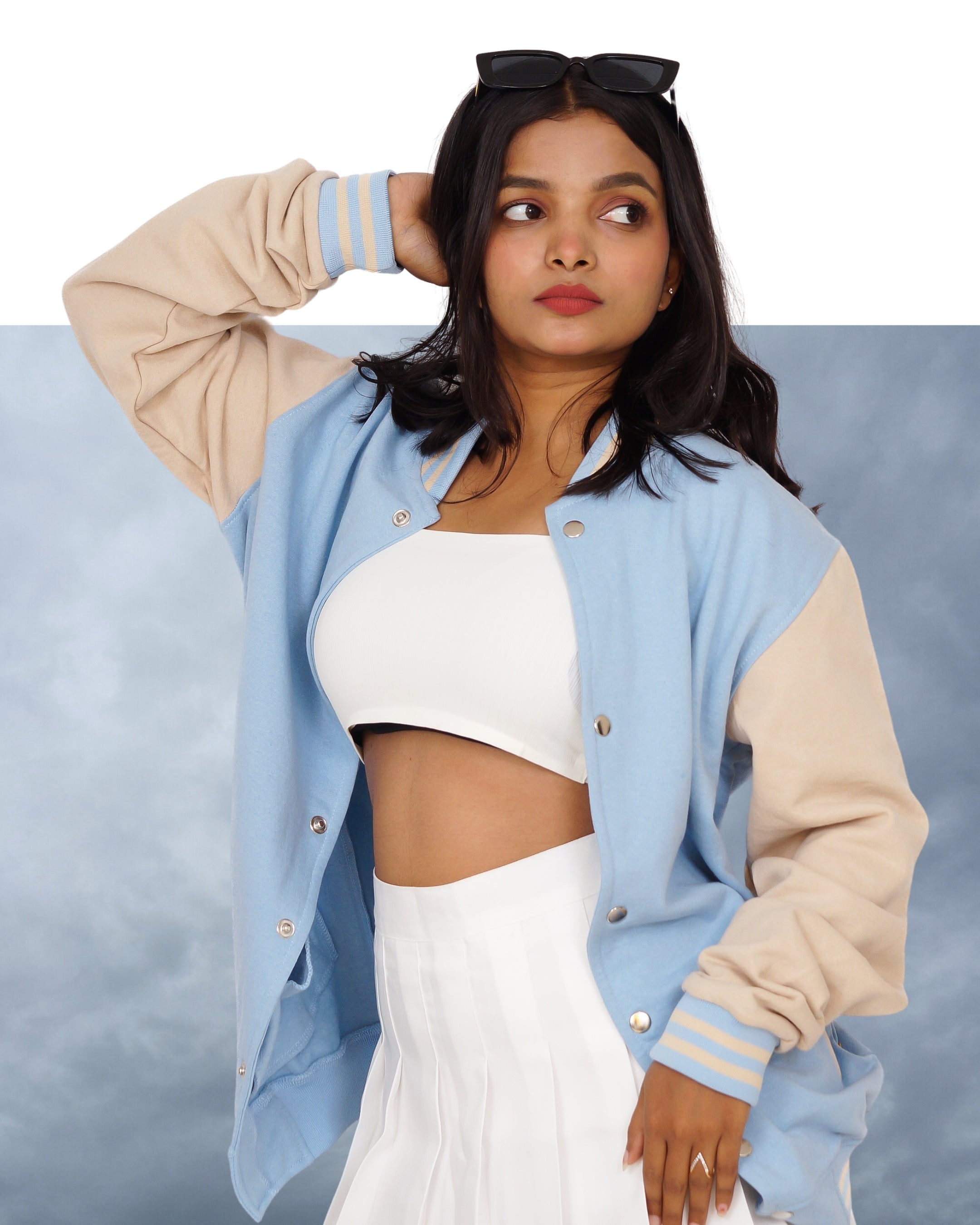 Woman wearing a light blue and beige varsity jacket with a white top and skirt on a gray background